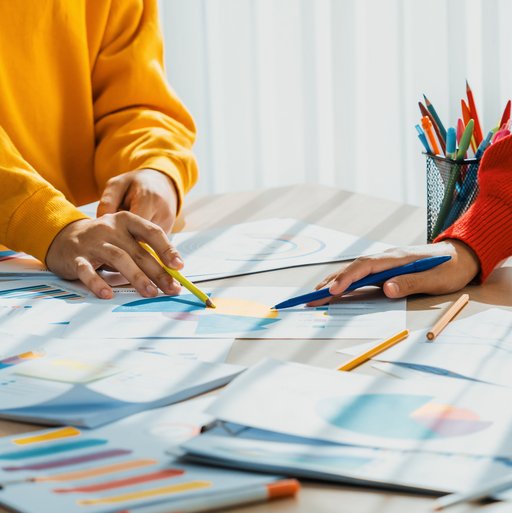 Drei Personen stehen an einem Tisch und arbeiten an bunten Grafiken auf Papier. Die Gesichter sind nicht sichtbar. 