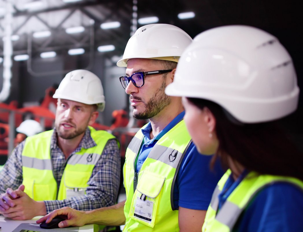 Drei Personen in einer Industriehalle stehen an einem Computer und schauen zusammen auf einen Bildschirm. 