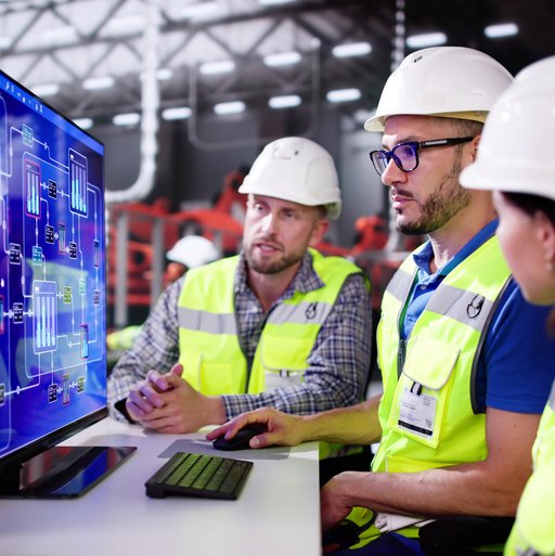 Drei Personen in einer Industriehalle stehen an einem Computer und schauen zusammen auf einen Bildschirm. 